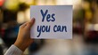 © HISTOCK - Woman holding a sign saying yes you can with blurred city background