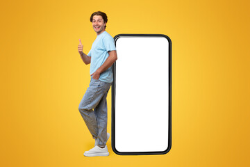 Cheerful young man standing leaning on big huge new cell phone with white empty screen and showing thumb up gesture, mockup, casual guy enjoying entertaining mobile app, orange studio background
