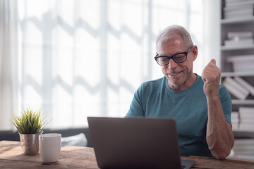 Wall Mural - Happy retired man raising his fist in celebration while using a laptop at home, enjoying the thrill of online success and good news in a cozy home office environment