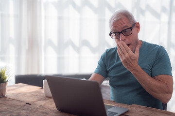 Wall Mural - Senior man with eyeglasses using laptop at home is shocked and surprised covering his mouth with hand while reading unbelievable news on computer screen