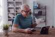 © MINAE - Happy retired man enjoying a cup of coffee while using smartphone and digital tablet at home, he is working remotely or attending an online meeting
