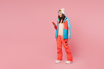 Wall Mural - Full body skier young woman wear warm blue padded jacket ski goggles mask spend weekend winter season in mountains hold in hand use mobile cell phone isolated on plain pink background. Hobby concept.