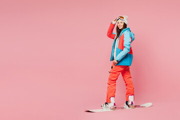 Full body side profile view young woman wear warm blue padded jacket ski goggles mask spend weekend winter season in mountains riding snowboard lean isolated on plain pink background. Hobby concept.