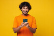 © ViDi Studio - Young smiling happy student Indian man he wear orange t-shirt casual clothes hold in hand use mobile cell phone chatting online isolated on plain yellow background studio portrait. Lifestyle concept.