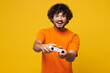 © ViDi Studio - Young shocked surprised happy Indian man he wear orange t-shirt casual clothes hold in hand play pc game with joystick console isolated on plain yellow background studio portrait. Lifestyle concept.