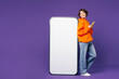 © ViDi Studio - Full body young smiling happy woman wear orange hoody casual clothes look at big huge blank screen mobile cell phone with area using smartphone isolated on plain purple background. Lifestyle concept.