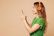 © ViDi Studio - Side view young woman she wear green t-shirt casual clothes listen to music in headphones hold in hand use mobile cell phone isolated on plain pastel light beige background studio. Lifestyle concept.