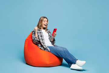 Wall Mural - Full body young woman wears checkered grey shirt casual clothes sit in bag chair hold in hand use mobile cell phone do winner gesture isolated on plain light blue cyan background. Lifestyle concept.