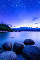 Wall Mural - Calm lake with rocks at night. Stars shine above mountains and water. Possible tourism