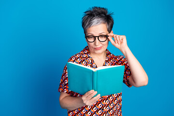 Wall Mural - Stylish senior woman with short grey hair and glasses enjoying leisure reading a book against a solid blue background