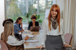 © bernardbodo - Red haired businesswoman posing in the glass office while her colleagues work