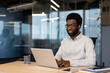 © Liubomir - Young successful businessman working on laptop inside office, man in light shirt smiling contentedly, typing on laptop keyboard at workplace.