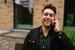 © dikushin - Portrait of cheerful stylish man enjoying pleasant phone conversation sitting on urban bench near modern urban building, smiling looking away. Concept of modern successful city lifestyle.