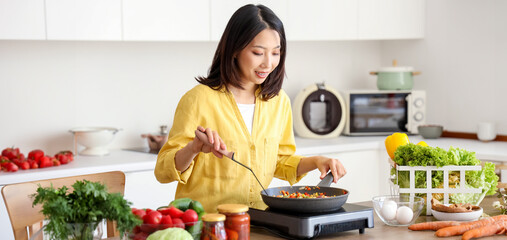 Wall Mural - Young Asian woman cooking different vegetables in frying pan on portable electric stove at kitchen