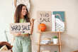© Pixel-Shot - Young African-American woman holding cardboard with text YARD SALE in room of unwanted stuff