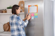 © Pixel-Shot - Young woman attaching sticky notes on fridge in modern kitchen