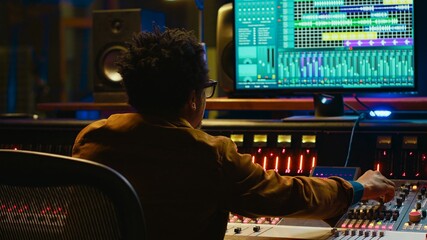Fototapete - Sound designer uses mixing console and managing audio tracks, twisting knobs on control desk for adjusting tunes and volume. Producer creates music in professional studio, technical gear. Camera A.