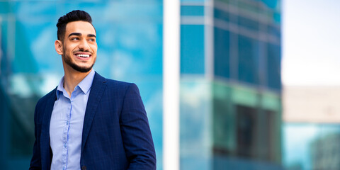 Wall Mural - Young bearded arab man manager in stylish suit holding hands in trousers pockets, standing by business center, looking at copy space and smiling, panorama. Career, job, work concept