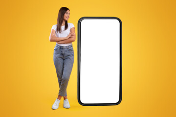 Wall Mural - cheerful smiling casual lady standing with crossed hands near giant big cell phone, empty white scre