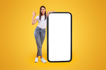 Wall Mural - Happy Woman Leaning On Big Smartphone With Blank White Screen And Gesturing Ok Sign, Cheerful Lady Recommending New App Or Website, Standing On Yellow Background, Mock Up Image, Full Body Length