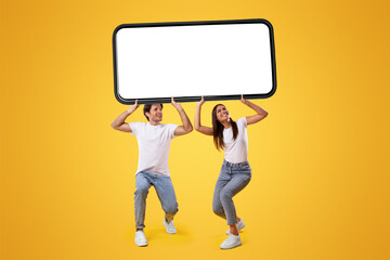 Wall Mural - millennial couple holding huge heavy smartphone with white empty screen on yellow orange studio back