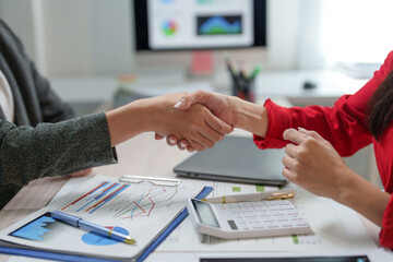 Wall Mural - Two businesswomen shaking hands after successfully reaching an agreement, analyzing financial charts, and using a calculator in a professional office environment