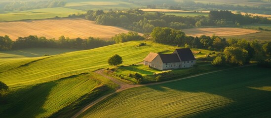 Poster - Aerial view of a picturesque rural farmhouse with a gray roof set amidst lush green fields and gentle rolling hills under a warm sunset glow
