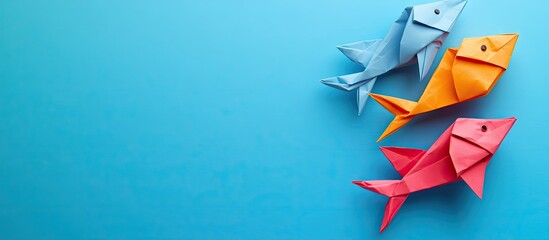Poster - Colorful origami fish in blue hues top view on a blue background featuring three fish in light blue, orange, and pink with ample copy space.