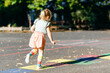 © Irina Schmidt - Cute little toddler girl playing hopscotch game drawn with colorful chalks on asphalt. Little active child jumping on playground outdoors on a sunny day. Summer activities for children.