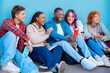 © alfons - A diverse group of five young students sit together on the floor, smiling and interacting on their smartphones against a blue background.
