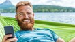 © A4719 - A man with a beard and a cheerful expression is lying in a lime green hammock, holding a smartphone. The background features a serene lake and