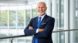 © Saowanee - confident senior businessman in suit stands with arms crossed, smiling in modern office setting. bright environment and his professional attire convey sense of success and elegance