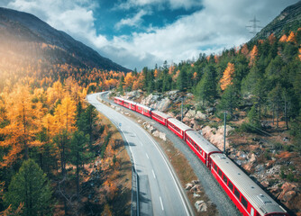 Wall Mural - aerial view of modern red train in alpine mountains, road, orange forest, sky with clouds at sunset 