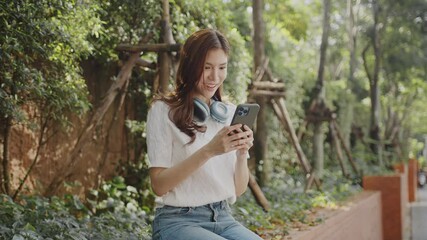 Wall Mural - Smiling woman enjoys outdoor technology while using her smartphone and wearing headphones in lush green park surrounded by nature.