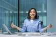 © Liubomir - Young joyful Asian woman meditating, resting and relaxing in lotus position. Woman smiling contentedly doing breathing exercises. Businesswoman inside office at workplace, calm and focused.