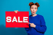 © deagreez - Young woman in blue sweater holding a vibrant sale sign against colorful background with cheerful expression