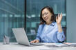 © Liubomir - Joyful and cheerful Asian woman talking to colleagues and partners using laptop for video call. Businesswoman at workplace inside office waving hand to interlocutor, greeting gesture.