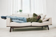 © SHOTPRIME STUDIO - Happy Black man relaxing on a modern sofa in a cozy white living room, wearing casual green sweater and blue jeans, showcasing comfort and joy