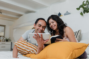 Wall Mural - Latin couple reading book and using tablet in bed at home in Mexico Latin America, hispanic people having fun