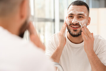 Wall Mural - Young man washing his face with soap near mirror in bathroom, closeup