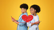 © Prostock-studio - A couple stands back to back against a vibrant yellow background, each holding a smartphone. They are sharing affectionate smiles while engaging with a messaging app on St.