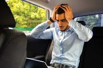 Wall Mural - Worried middle eastern businessman using laptop while going to business meeting by car, sitting on auto back seat and touching his head with both hands, having troubles at job, copy space