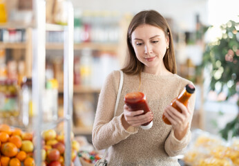 Wall Mural - Young woman buyer choosing ketchup or chili sauce in bottle in grocery store