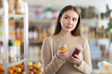 Wall Mural - Young woman buyer chooses red and yellow smoothie in bottle in grocery store