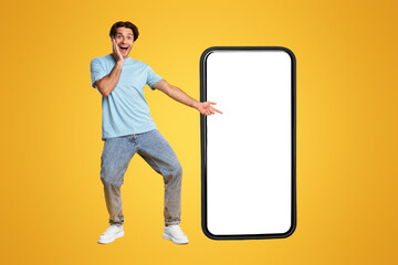 Wall Mural - Great Mobile Offer. Excited Guy Pointing Finger At Big White Smart Phone Screen, Emotionally Reacting To New App, Overjoyed Young Male Standing Isolated Over Orange Studio Background, Mock Up