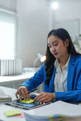 Wall Mural - Businesswoman in a blue suit working diligently at her desk, analyzing financial reports on a tablet while jotting down important notes on sticky notes for effective planning