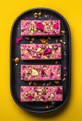 Wall Mural - Colorful dessert bars with rose petals and nuts on black plate against bright yellow background, perfect for festive occasions and culinary photography