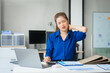 © Phushutter - Asian businesswoman sits at her ergonomic desk, working on a laptop and mobile phone. Her posture ensures minimal strain, addressing neck pain, back pain, and preventing repetitive strain injuries.