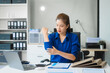 © Phushutter - Asian businesswoman sits at her ergonomic desk, working on a laptop and mobile phone. Her posture ensures minimal strain, addressing neck pain, back pain, and preventing repetitive strain injuries.