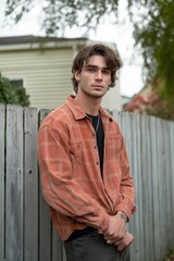 Wall Mural - A man in an orange shirt stands in front of a wooden fence. He is wearing a black shirt and a necklace